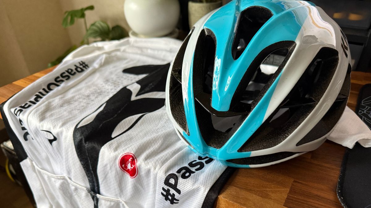 室内の木製テーブルに準備された水色と白のKASK（カスク）製ロードバイク用ヘルメットと、折りたたまれたカステリのサイクルジャージ。初心者が休日のソロライドに出発する前の、安全で快適な装備を整える心弾む準備時間の風景。