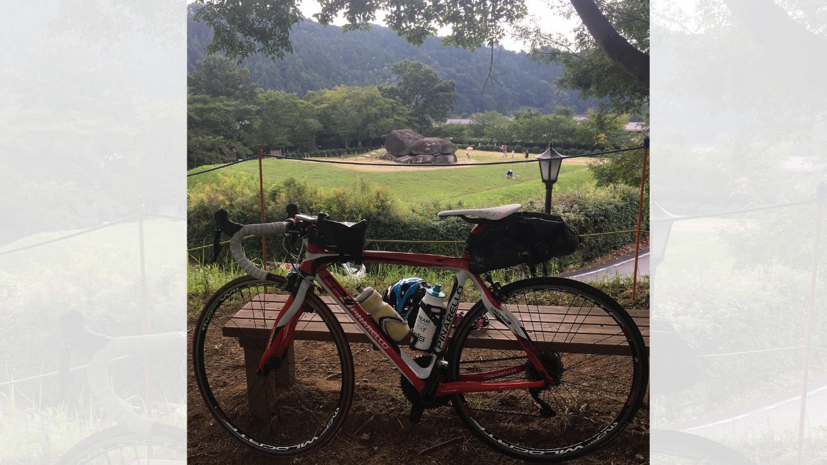 緑豊かな飛鳥の風景と石舞台古墳を背景に、木製ベンチで休憩中の赤と白のロードバイク(ピナレロ)。ダブルボトルや大きめのサドルバッグなど、初心者が安全に長距離のソロライドを楽しむための装備を備えています。歴史ある名所を巡る自転車旅の風景。