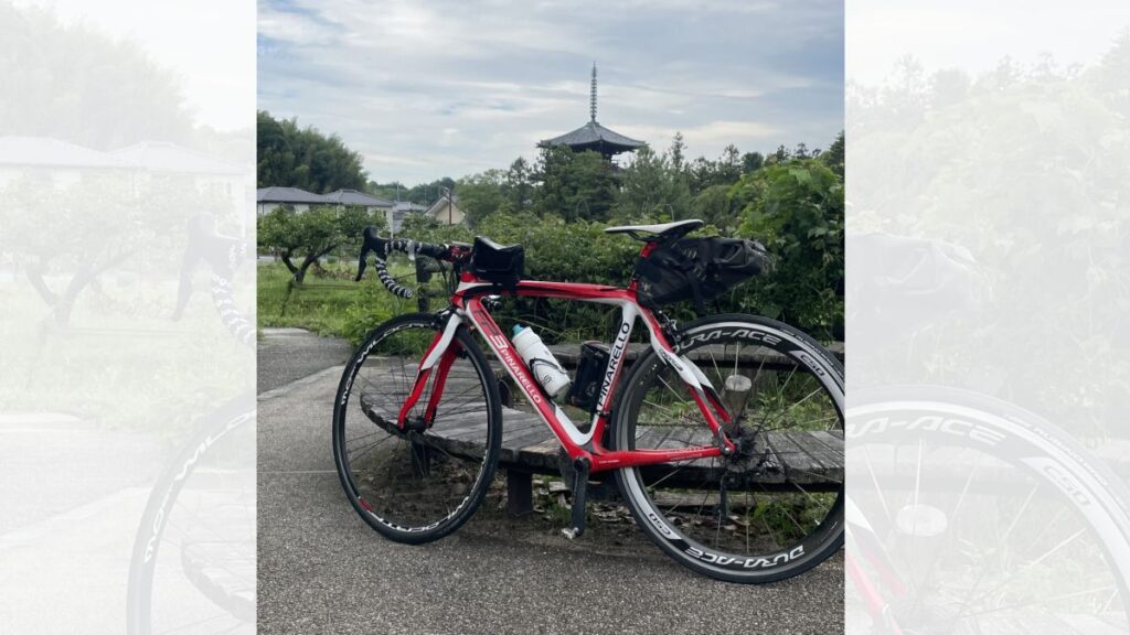 遠くに斑鳩の法輪寺・三重塔を望む緑豊かな小道で、木製のベンチに立てかけられた赤と白のロードバイクの写真。歴史ある風景を楽しみながら走る、初心者にもおすすめのサイクリングルートを伝える画像。