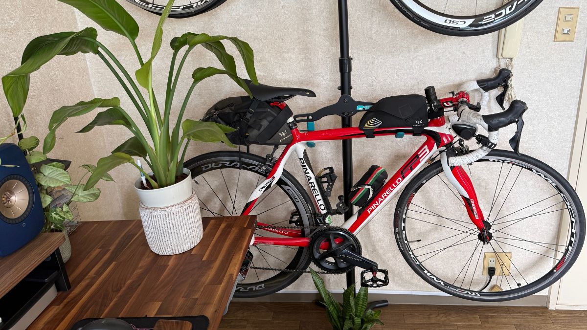 室内用上下2段式バイクスタンドの下段に収納された赤と白のロードバイク（ピナレロ）。デスクや観葉植物の隣に省スペースで収まる様子。初心者が悩みがちな室内保管において、縦置きスタンドを活用し生活空間とスッキリ両立させるための参考レイアウト。