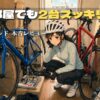 荷物と2台のロードバイクで窮屈になった部屋で、収納場所に悩む女性サイクリストのイラスト。「狭い部屋でも2台スッキリ！上下2段スタンド本音レビュー」の文字が添えられ、室内保管の悩みを解決するアイテムを初心者に分かりやすく紹介するアイキャッチ。