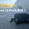道路脇に置かれた黒いAPIDURAの1Lトップチューブバッグ。「iPhone 16 Proも余裕！本音レビュー」の文字がある。
