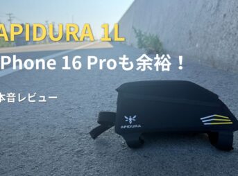 道路脇に置かれた黒いAPIDURAの1Lトップチューブバッグ。「iPhone 16 Proも余裕！本音レビュー」の文字がある。