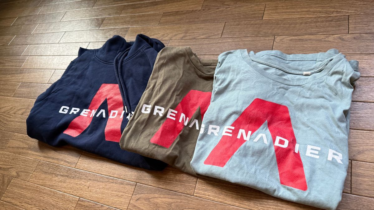 床に並べられたINEOS Grenadiersのロゴ入りパーカーとTシャツ2枚。「推しチームを、普段着に。」の文字がある。