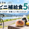 川沿いのベンチにロードバイクを停め、あんぱんやおにぎり、バナナなどの補給食を並べた休憩風景。初心者がハンガーノックを防ぎ、手軽にエネルギーチャージできる定番アイテムを紹介する「ロードバイクのコンビニ補給食5選」のアイキャッチ。