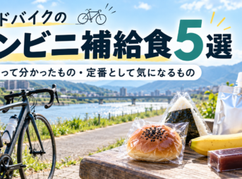 川沿いのベンチにロードバイクを停め、あんぱんやおにぎり、バナナなどの補給食を並べた休憩風景。初心者がハンガーノックを防ぎ、手軽にエネルギーチャージできる定番アイテムを紹介する「ロードバイクのコンビニ補給食5選」のアイキャッチ。