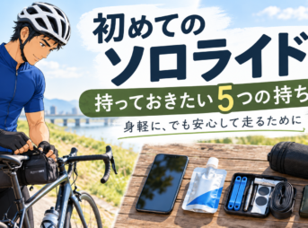 川沿いでロードバイクのサドルバッグを準備する男性サイクリストと、テーブルに並んだスマートフォン、補給食、パンク修理キットなどの必須アイテム。初心者が初めてのソロライドに安心して出かけるための、身軽で安全な持ち物5選を解説するアイキャッチ。